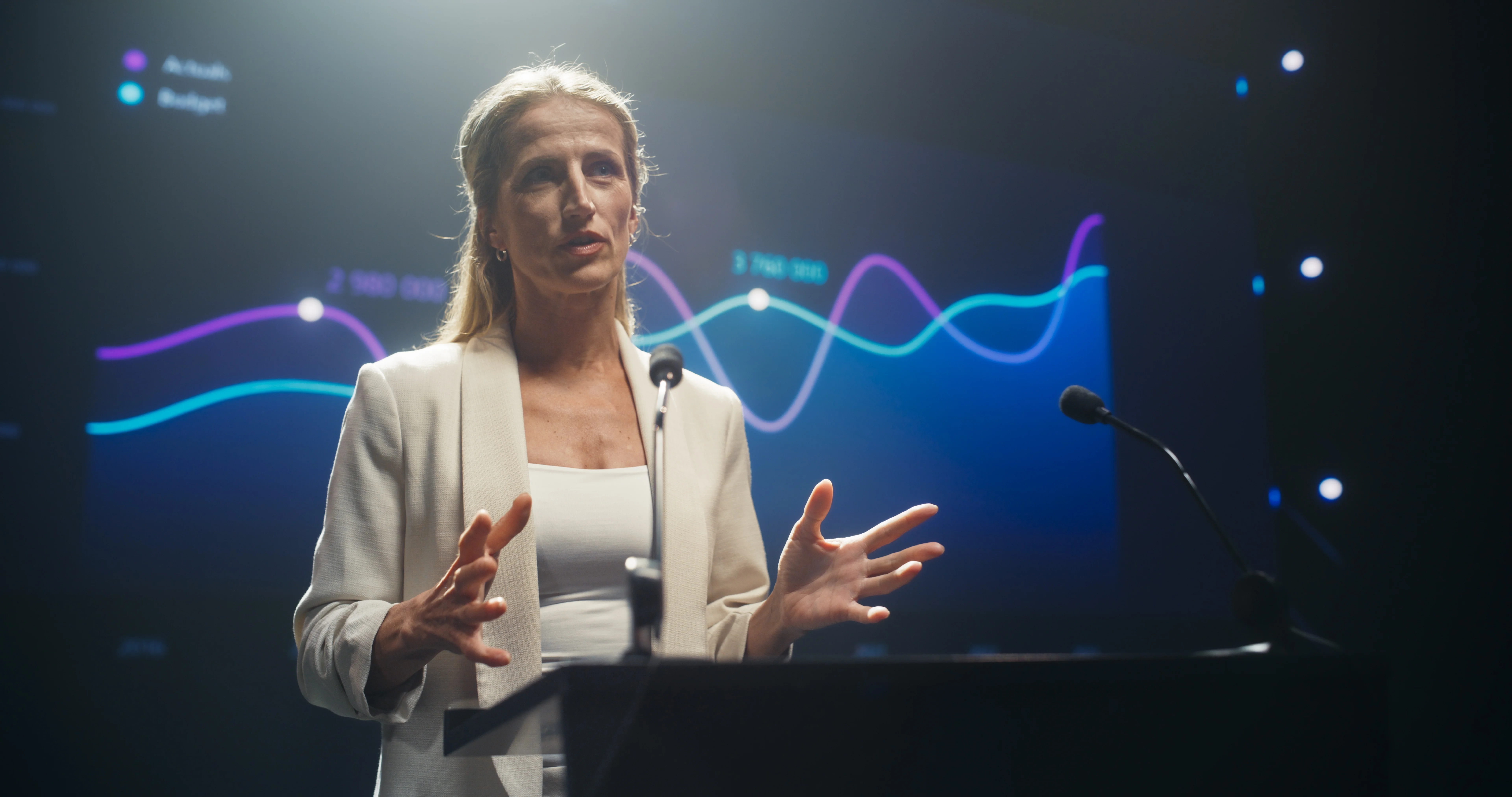 Woman in white blazer presenting data with colorful line graphs on a large screen behind her.
