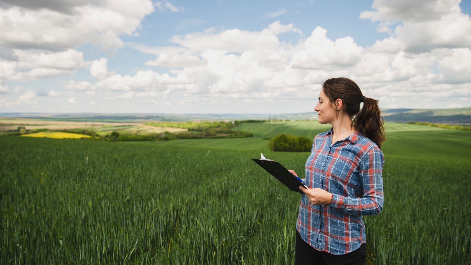 Woman in a plaid shirt holding a clipboard and standing in a lush green agricultural field under a partly cloudy sky.