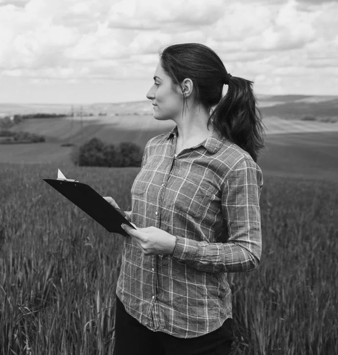 Woman with dark hair in a ponytail wearing a plaid shirt holding a clipboard and standing in a field with rolling hills in the background.