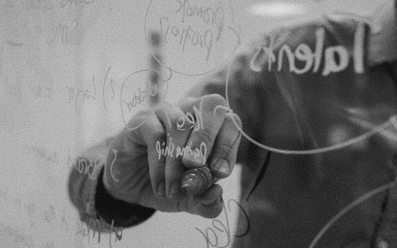 Person writing or drawing on a transparent glass board with a marker, showing handwritten words and diagrams.