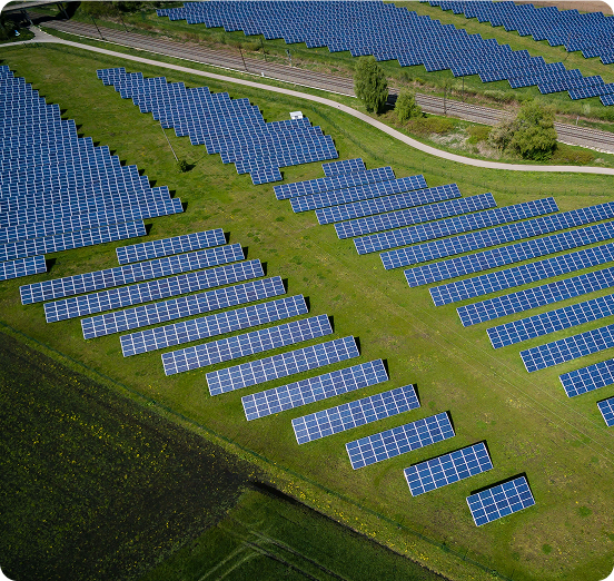 Aerial view of a solar farm with multiple rows of blue solar panels installed on green grass beside a narrow road and railway tracks.