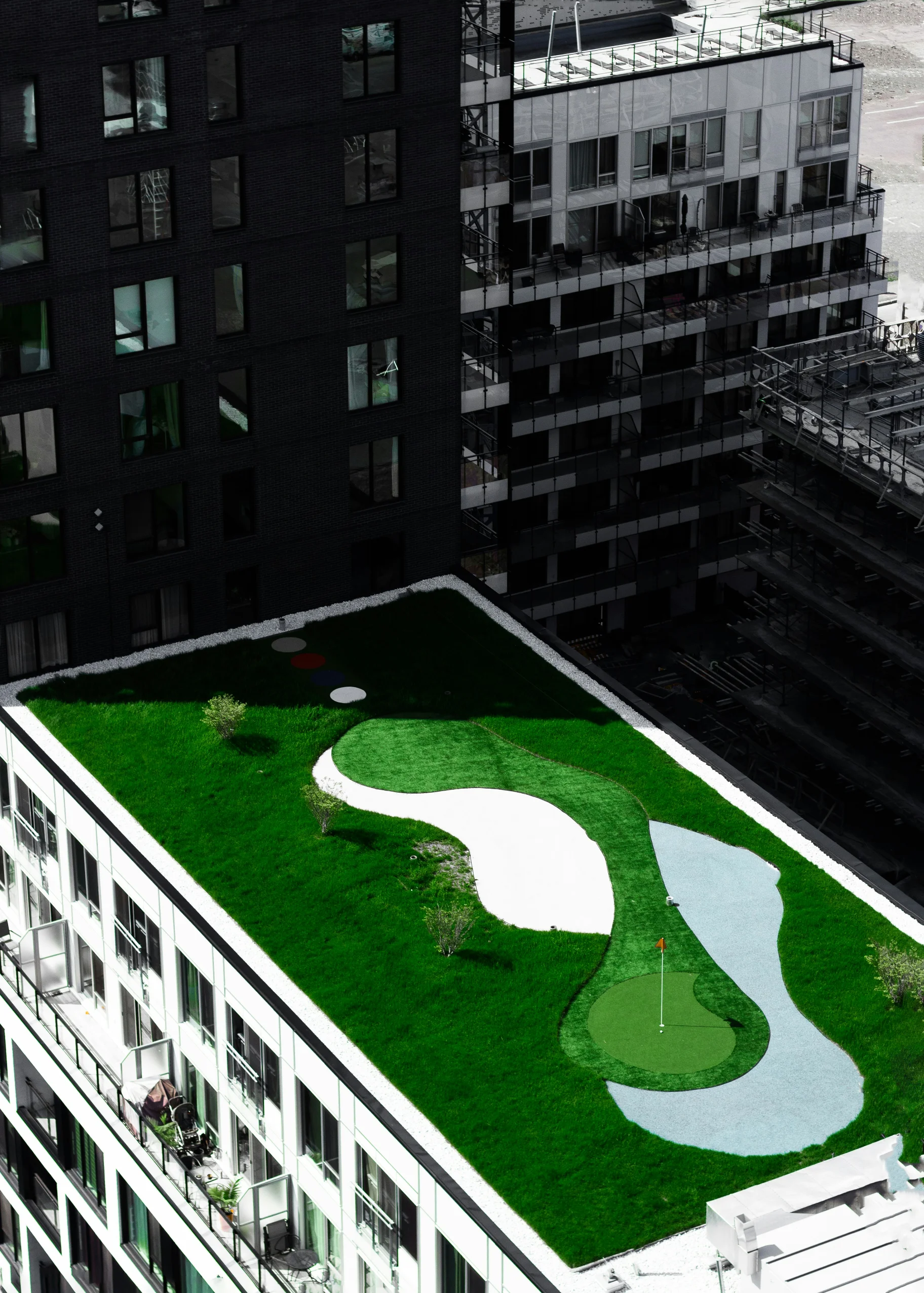 Rooftop green space with a mini golf putting green, sand bunker, and artificial water hazard on top of a white building in an urban area.