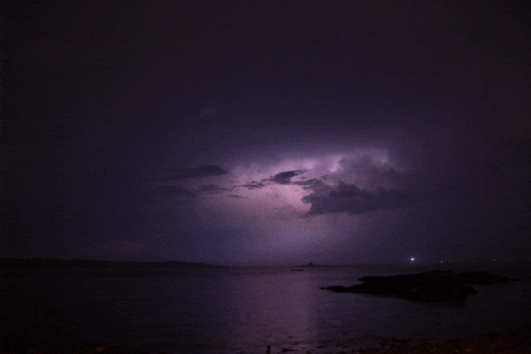 Lightning illuminates dark storm clouds over a calm sea with rocky shore at night.