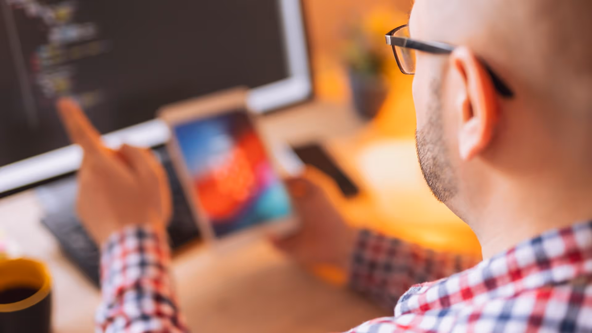 Bald man wearing glasses and plaid shirt pointing at a computer screen while holding a tablet.