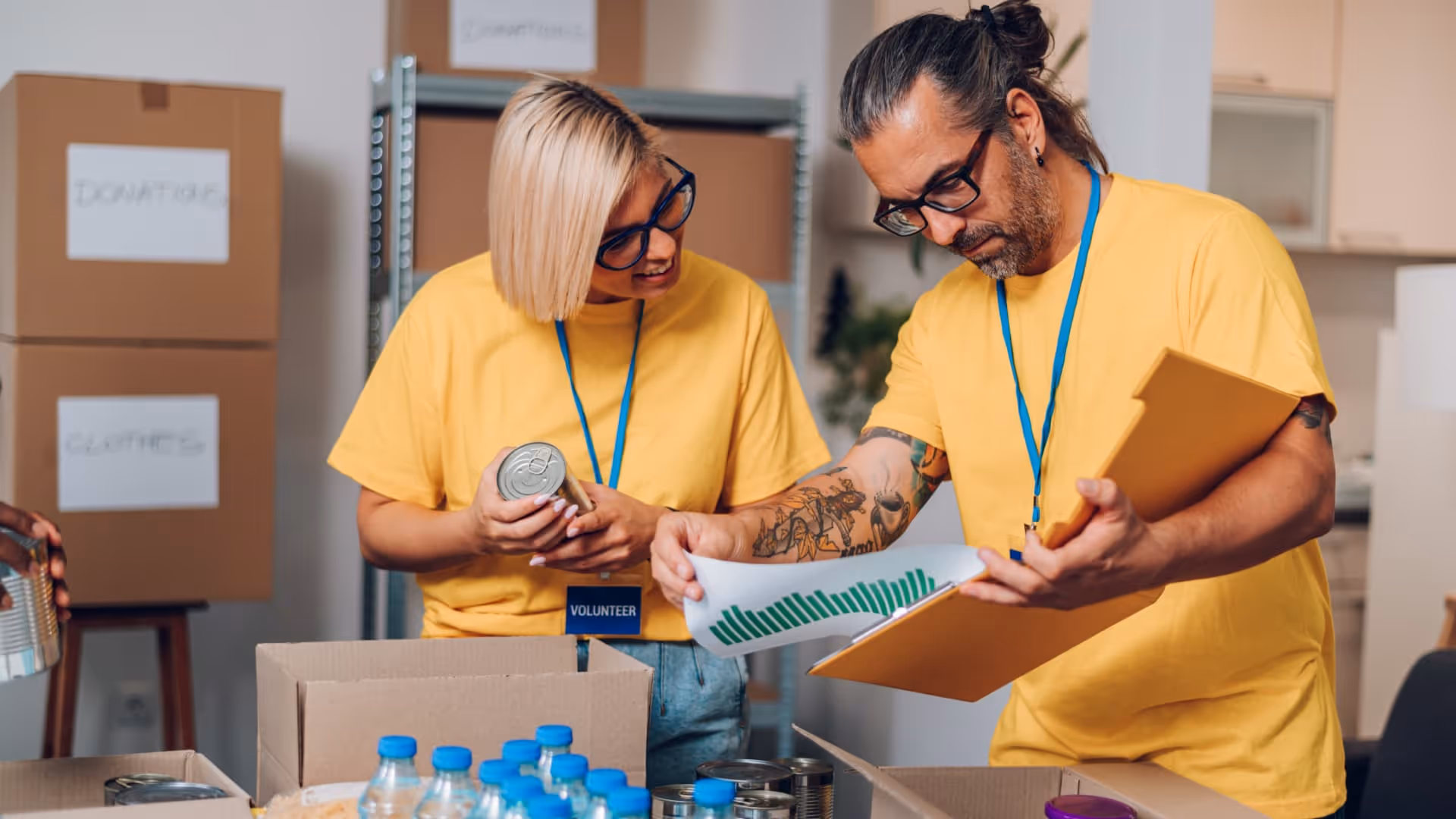 Two volunteers in yellow shirts organizing canned goods and bottled water in a community charity setting.