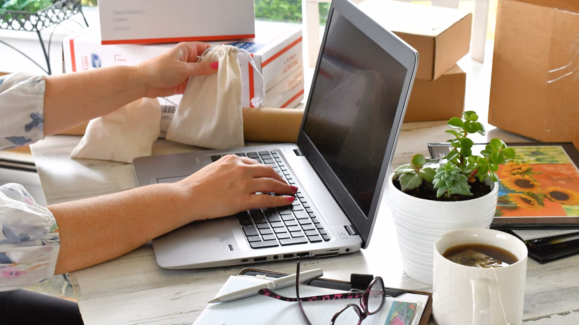 Person using a laptop while holding a small drawstring bag with boxes and a coffee cup nearby.