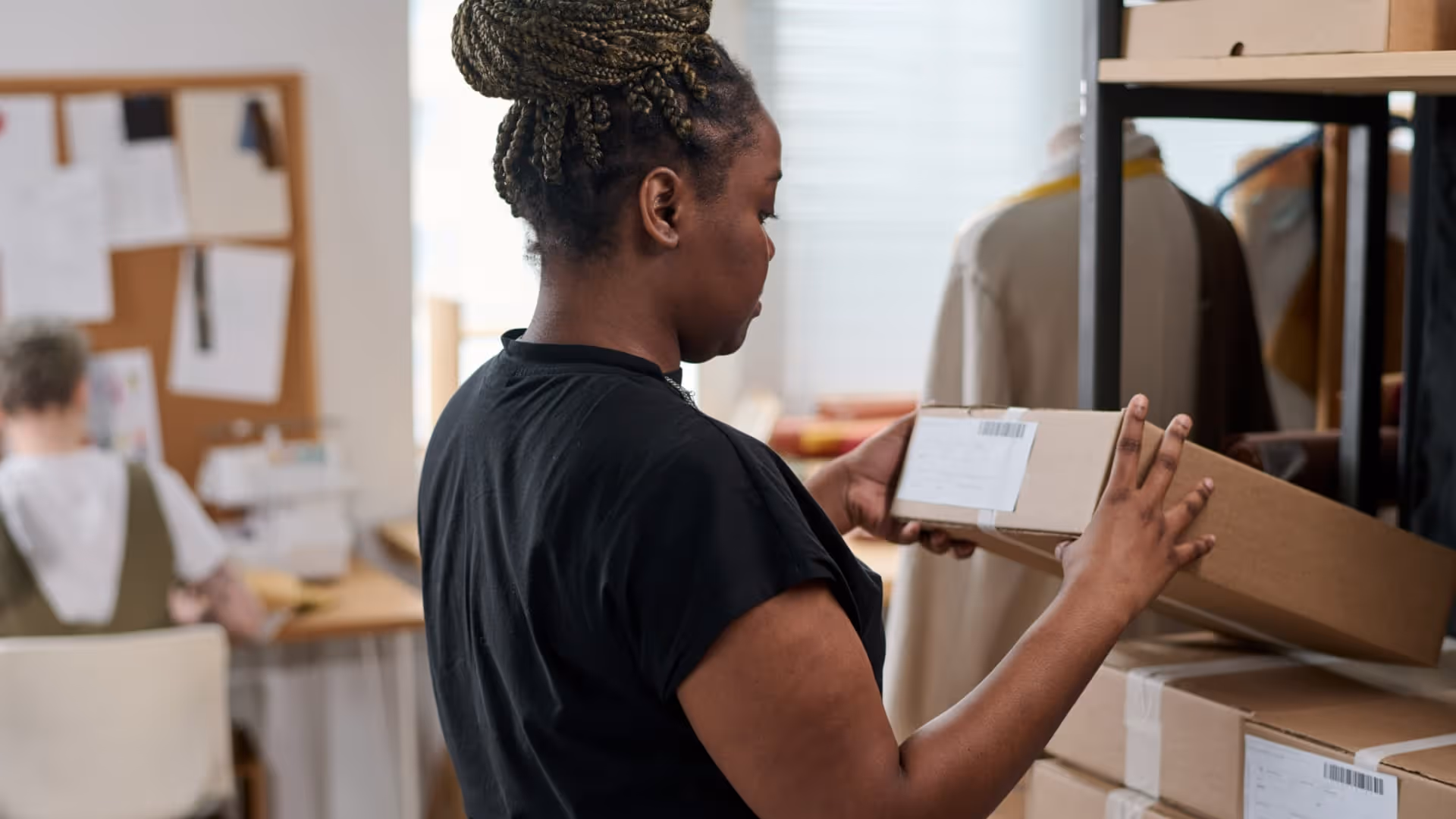 Woman with braided hair checking address label on a shipping box in a workspace.