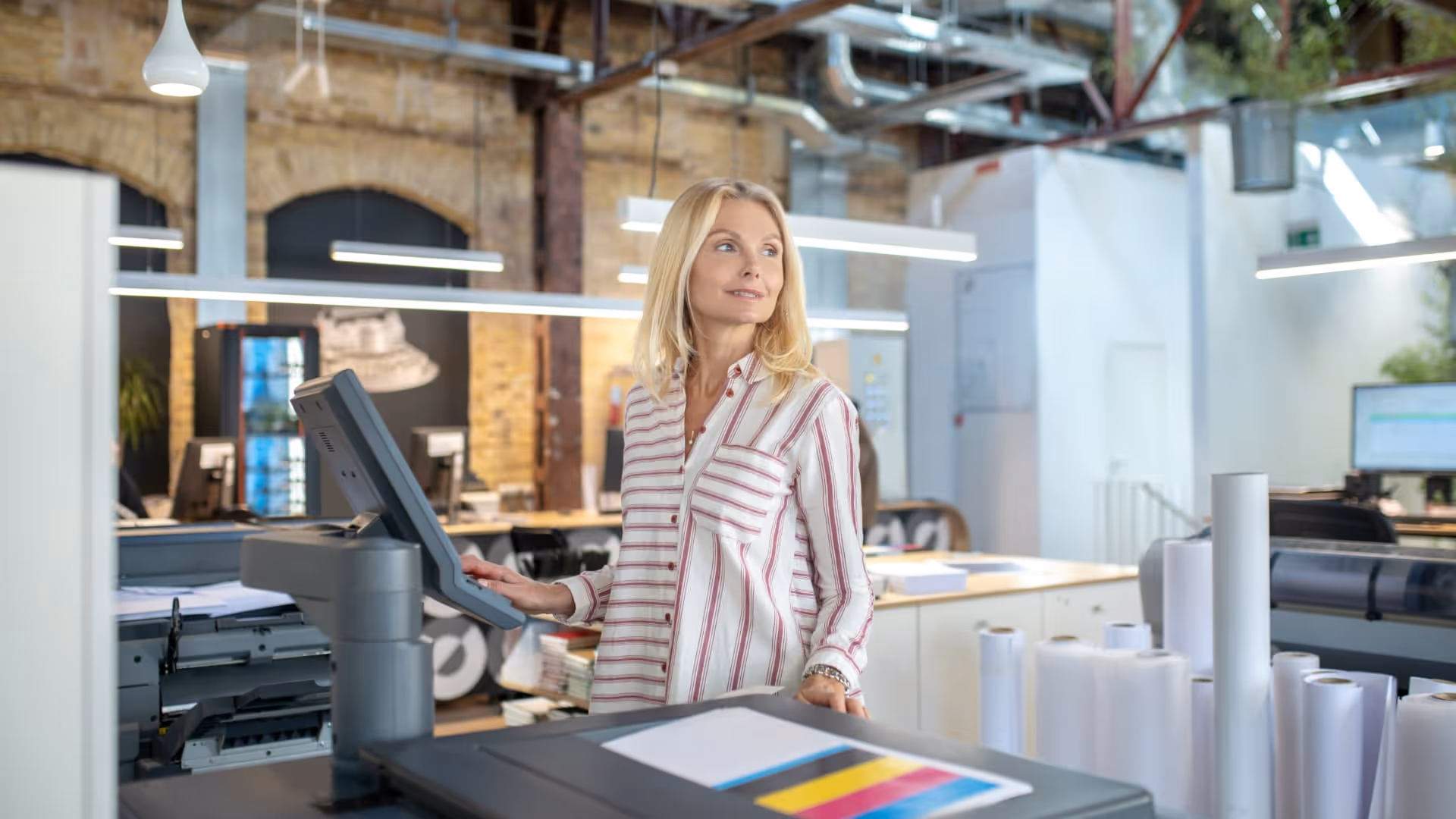 Blonde woman adjusting settings on a large printer in a modern office with paper rolls nearby.