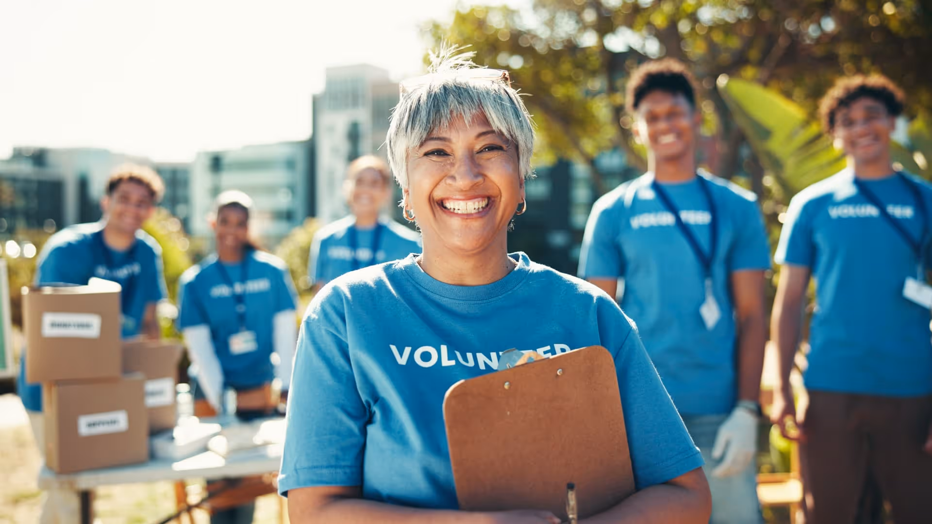 Smiling woman volunteer holding a clipboard with volunteers and donation boxes in the background.
