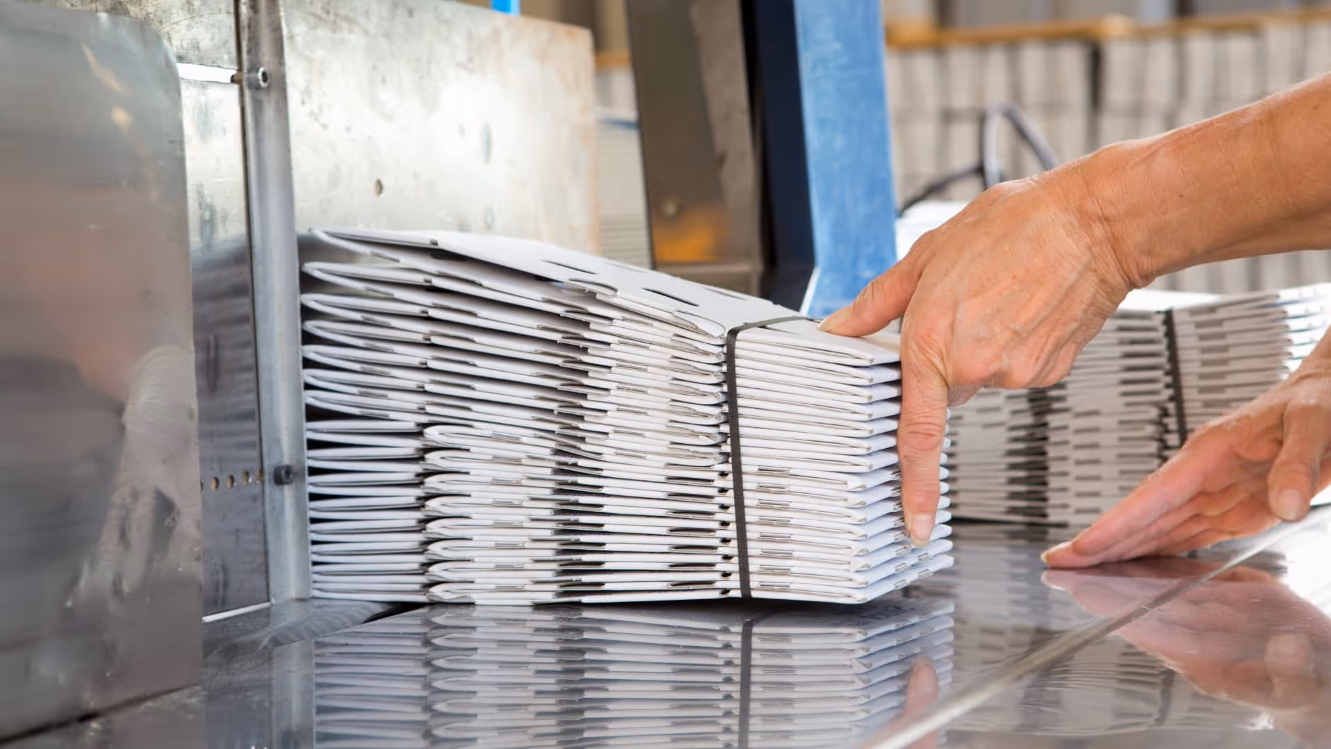 Hands stacking a bundle of folded white cardboard boxes tied with a black strap on a reflective metal surface.