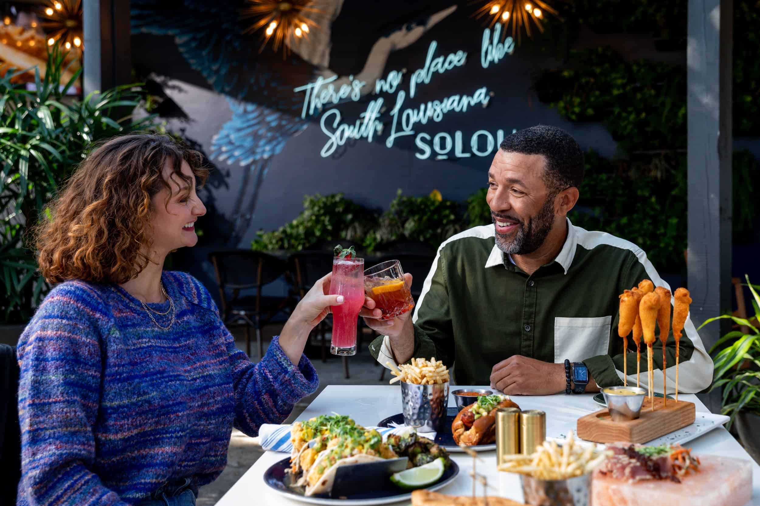 A couple toasting over a meal beneath a neon sign reading There's No Place Like South Louisiana