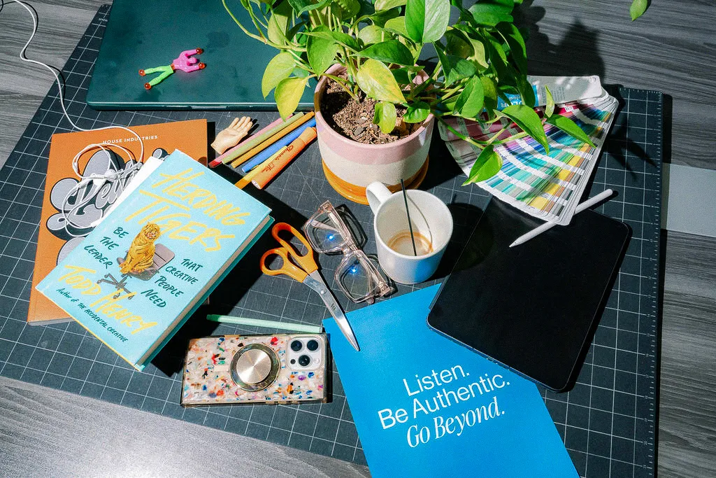 An overhead view of a desk with design books, a plant, scissors and a blue card reading Listen. Be Authentic. Go Beyond.