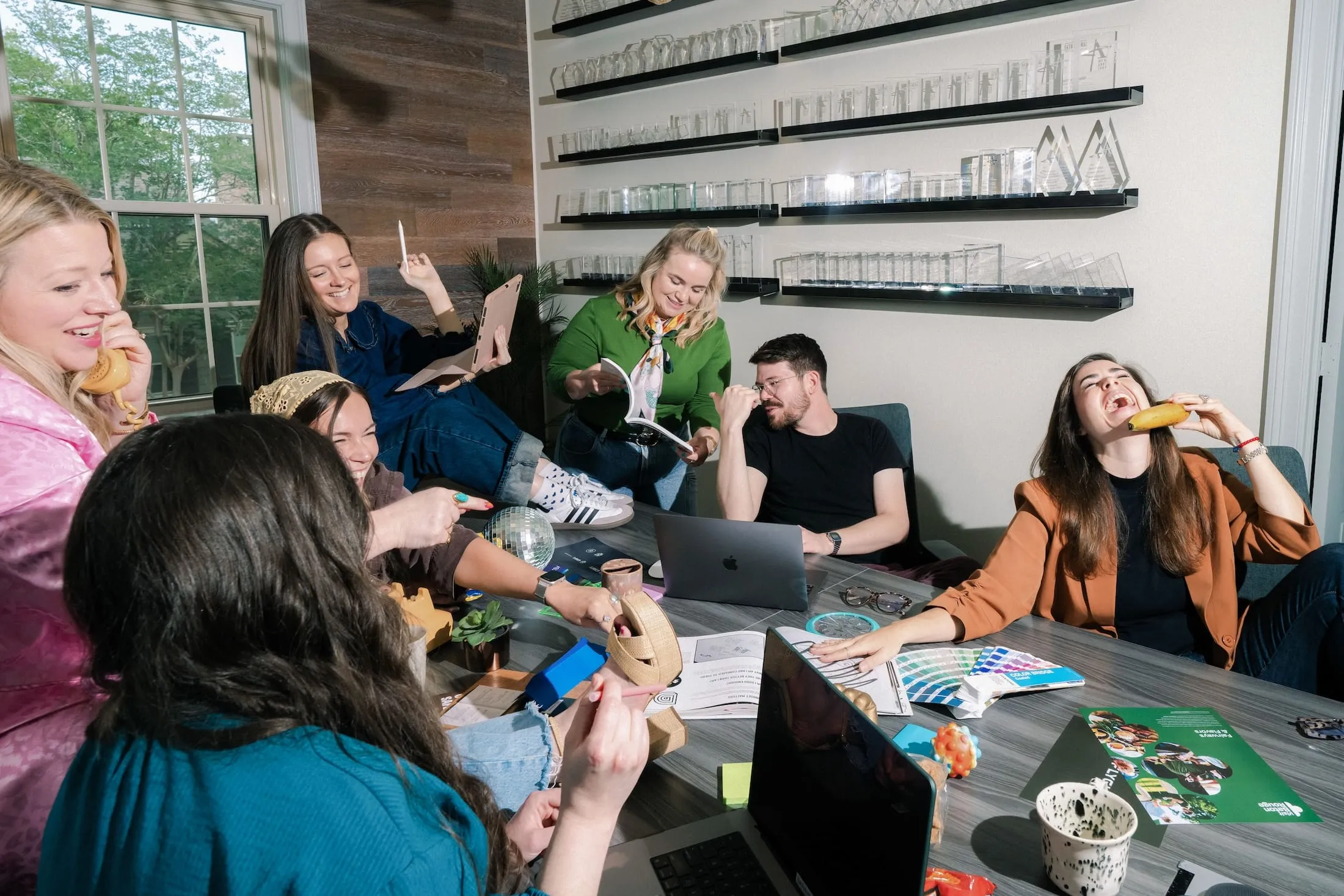 Team members gathered around a table laughing, several holding bananas to their ears like phones