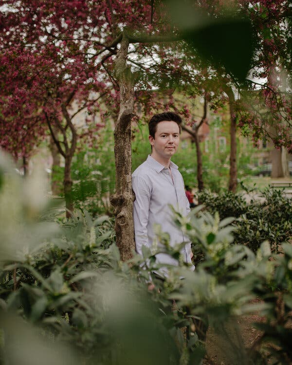 Billy Casagrande stands against the narrow trunk of a tree in a lush Brooklyn park.