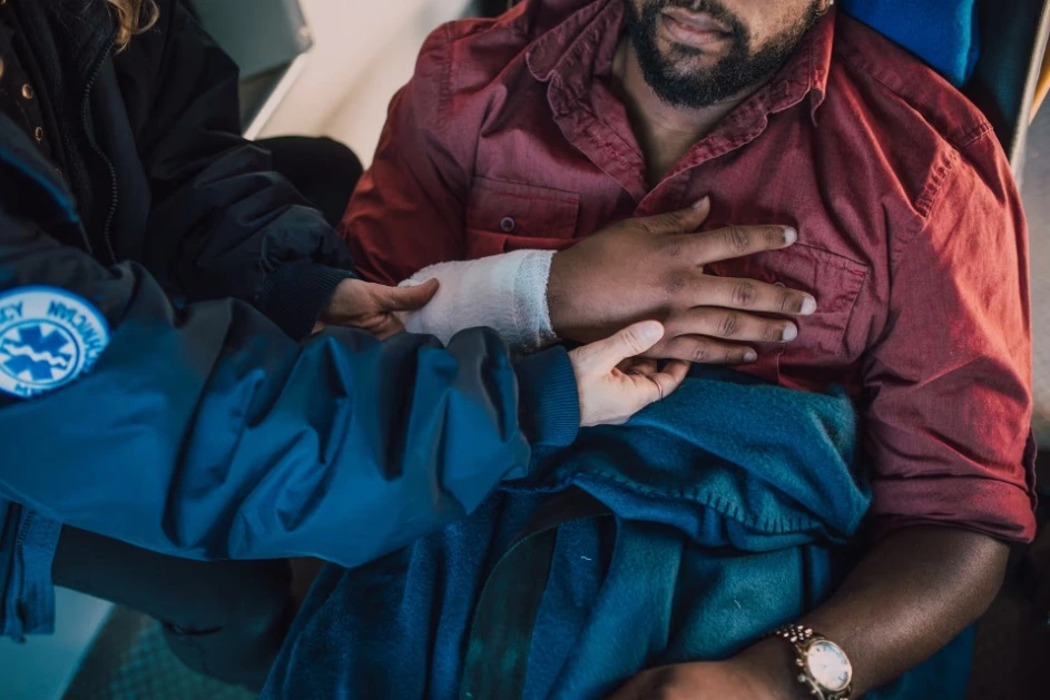 A paramedic in a dark blue jacket with an EMS patch applying a white bandage or checking an injury on a male patient's forearm while he sits wrapped in a blue blanket.