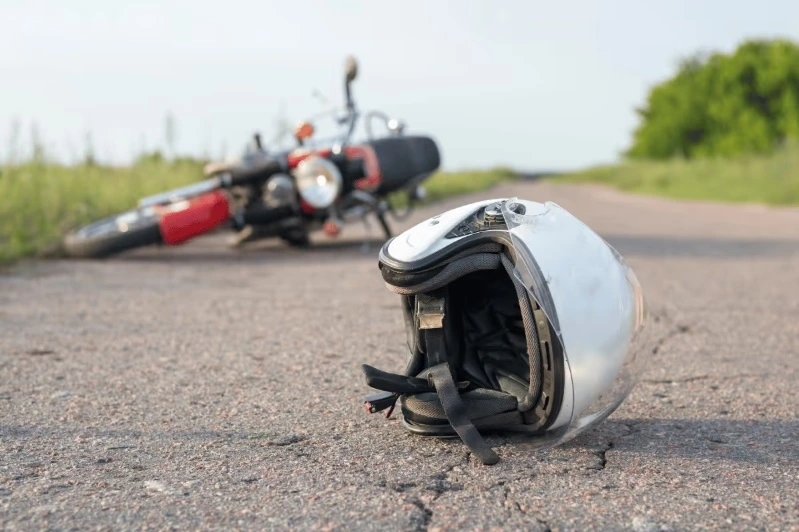 A white motorcycle helmet resting on cracked asphalt in the foreground, with an overturned motorcycle lying on the roadside in the blurred background, suggesting a recent accident.