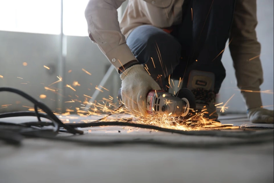 Low-angle close-up of a construction worker kneeling and wearing gloves and boots, using an angle grinder tool to cut or grind metal on the floor, producing a shower of bright orange sparks.