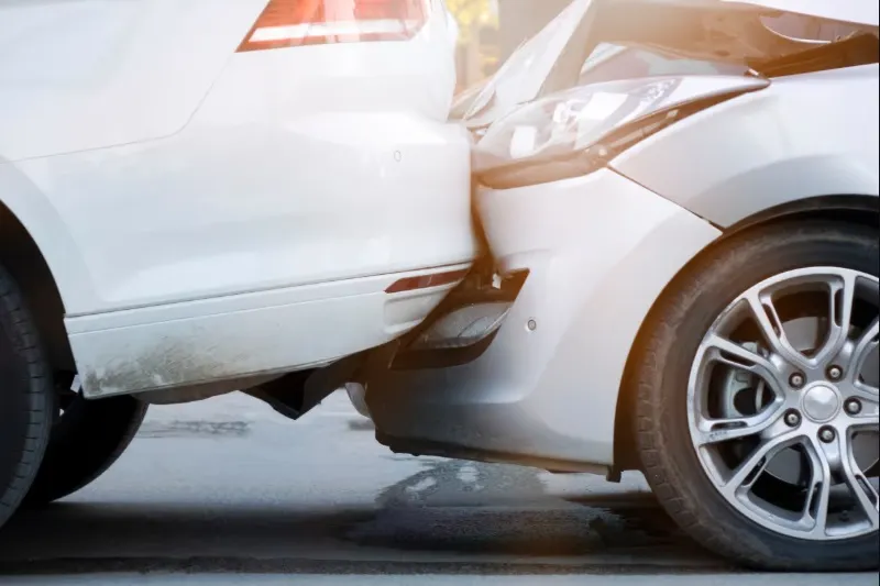 Close-up image of a rear-end car accident, showing the front bumper and headlight of one white car impacting the rear bumper of a second white car.