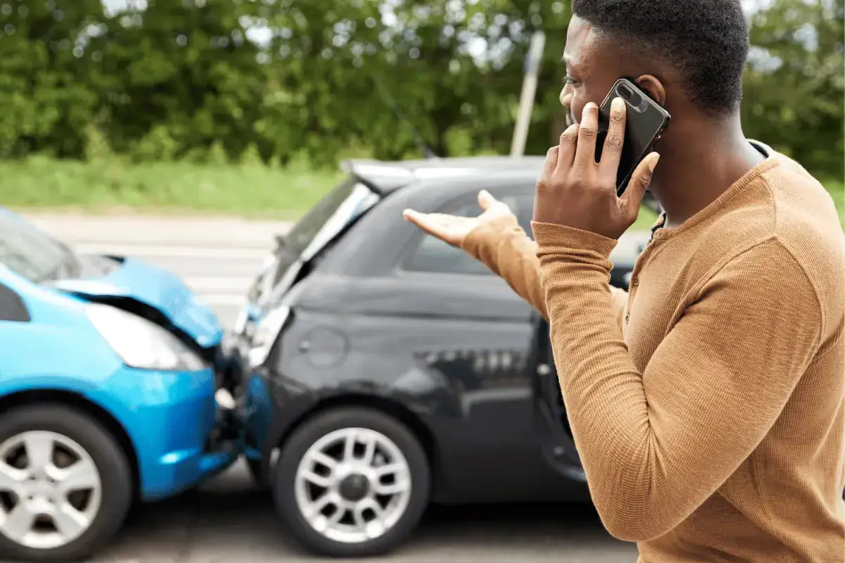 A distressed male driver is standing on the street, speaking on his mobile phone after a rear-end collision between a blue hatchback and a black car behind it. Damage to the vehicles is visible in the background.