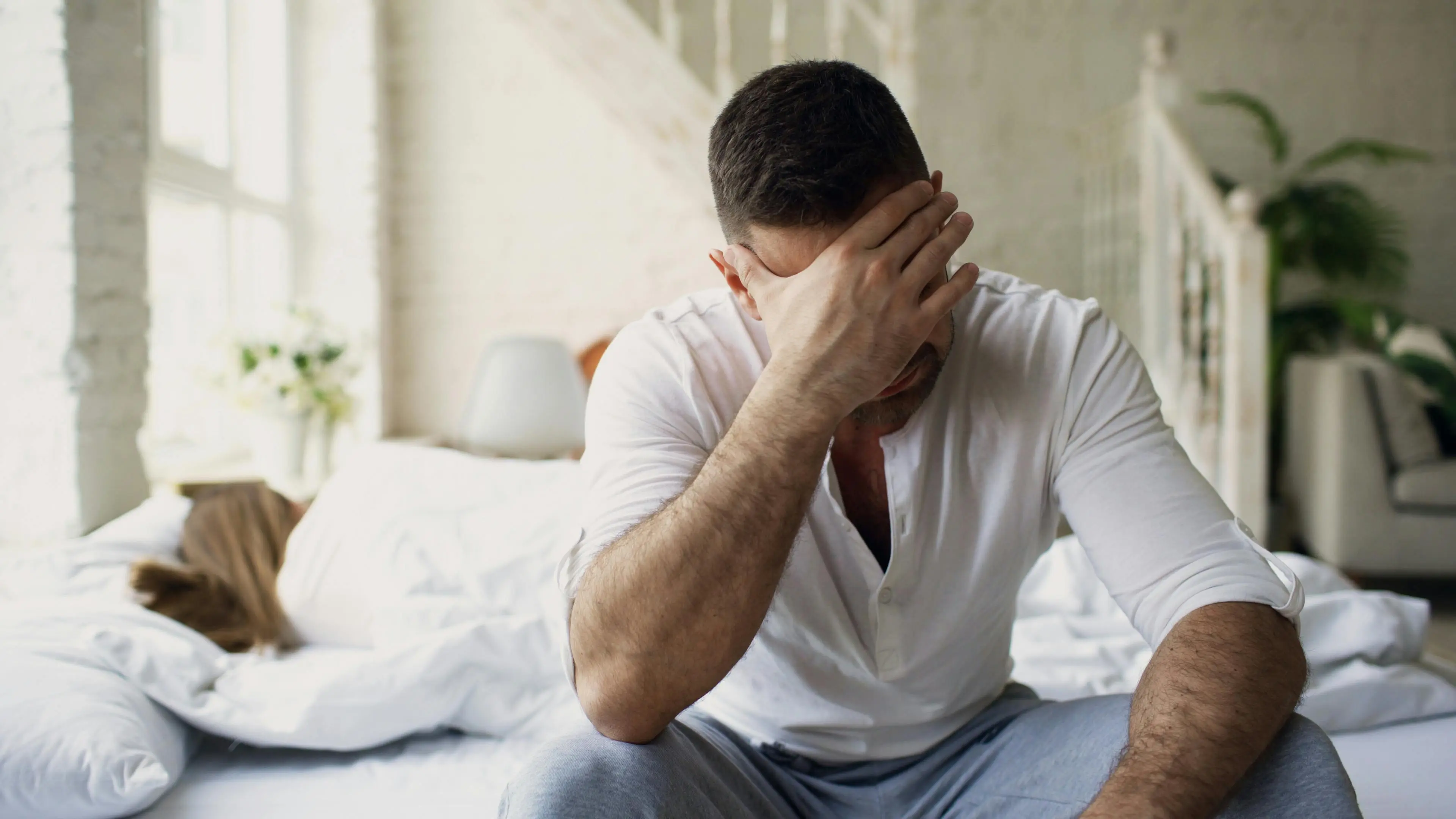 Man sitting on a bed with his hand covering his face, looking stressed, while a woman lies in bed behind him.