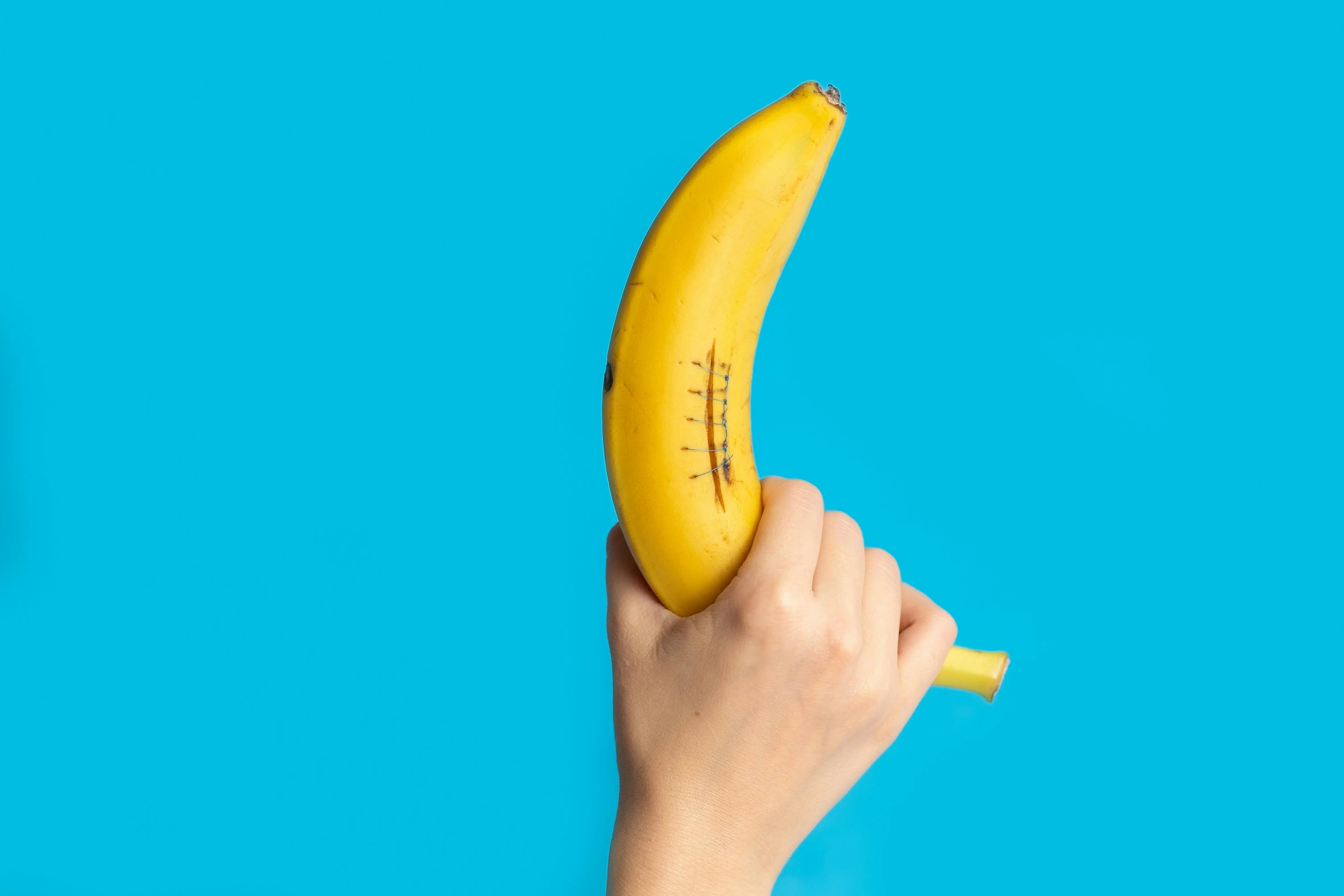 Hand holding a banana with drawn stitches on its peel against a blue background.