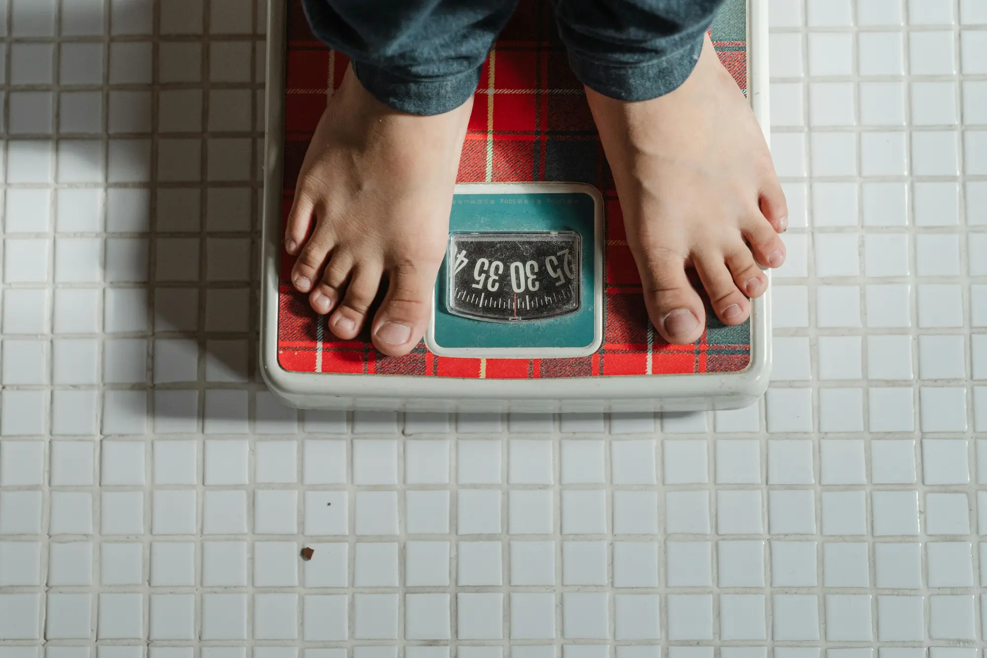 Bare feet standing on a red plaid patterned analog weighing scale on white tiled floor.