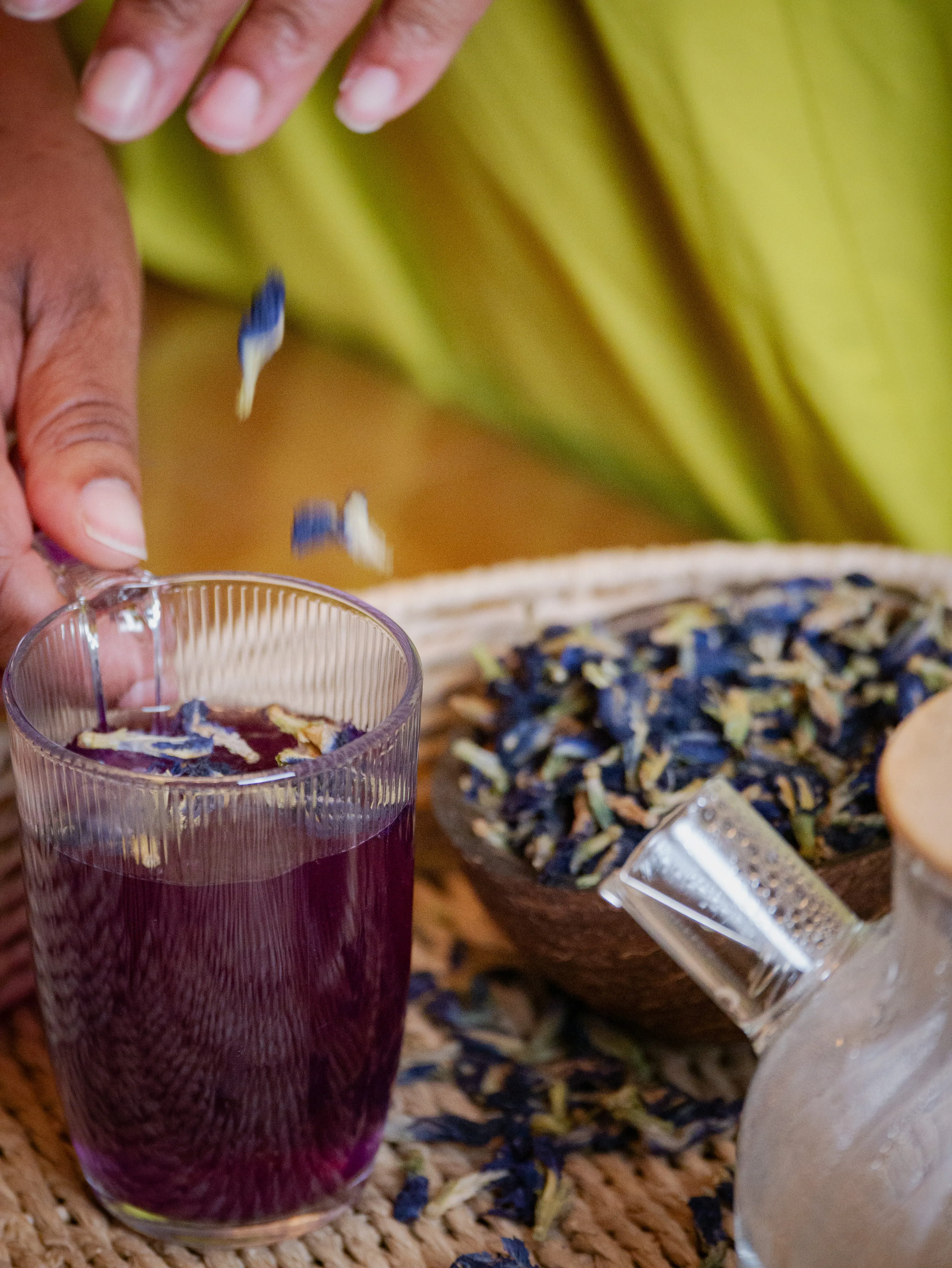 Une main verse des fleurs séchées bleu violet dans un verre transparent de tisane.