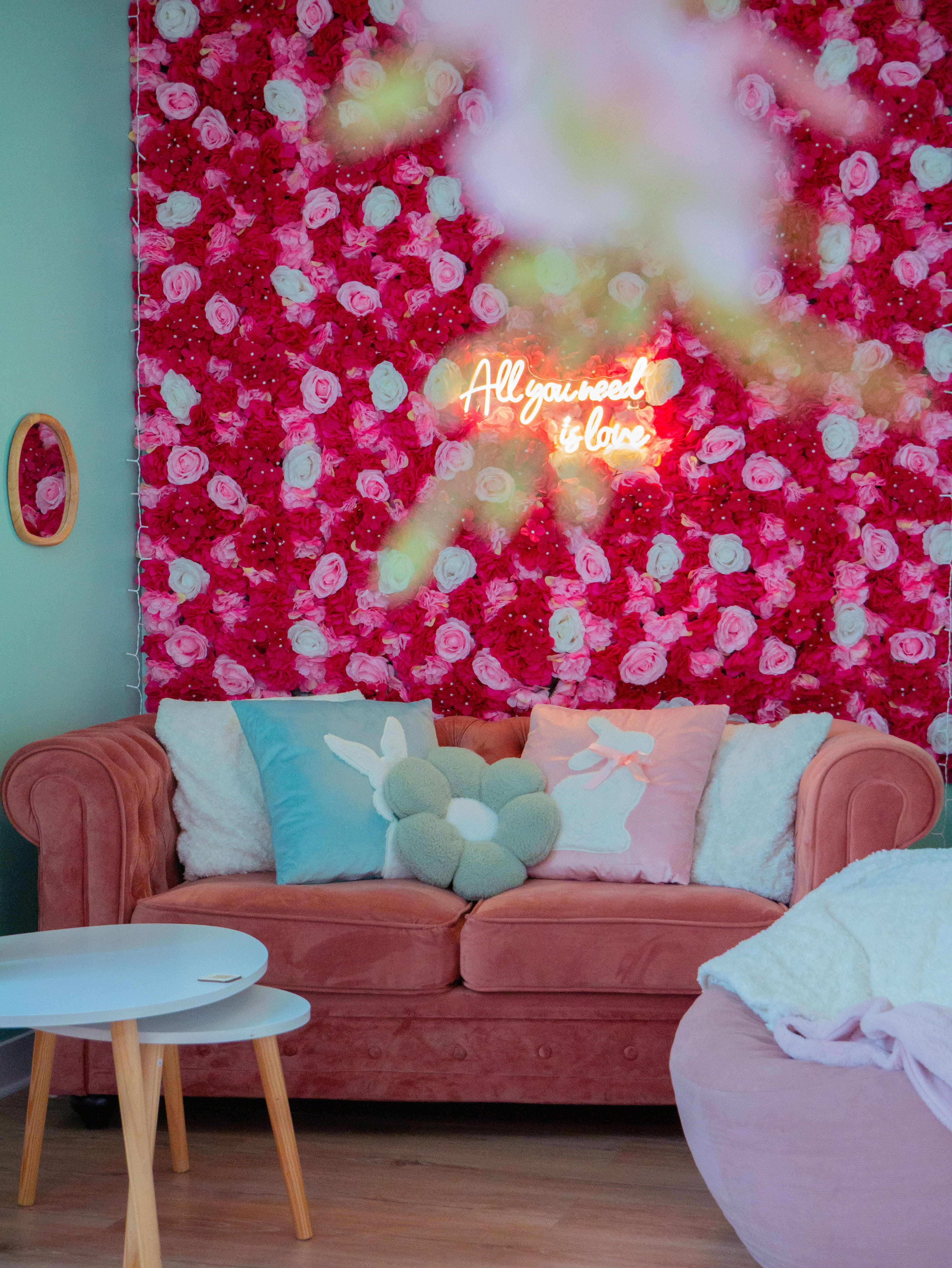 Canapé rose avec coussins pastel devant un mur de fleurs et un néon disant « All you need is love ».