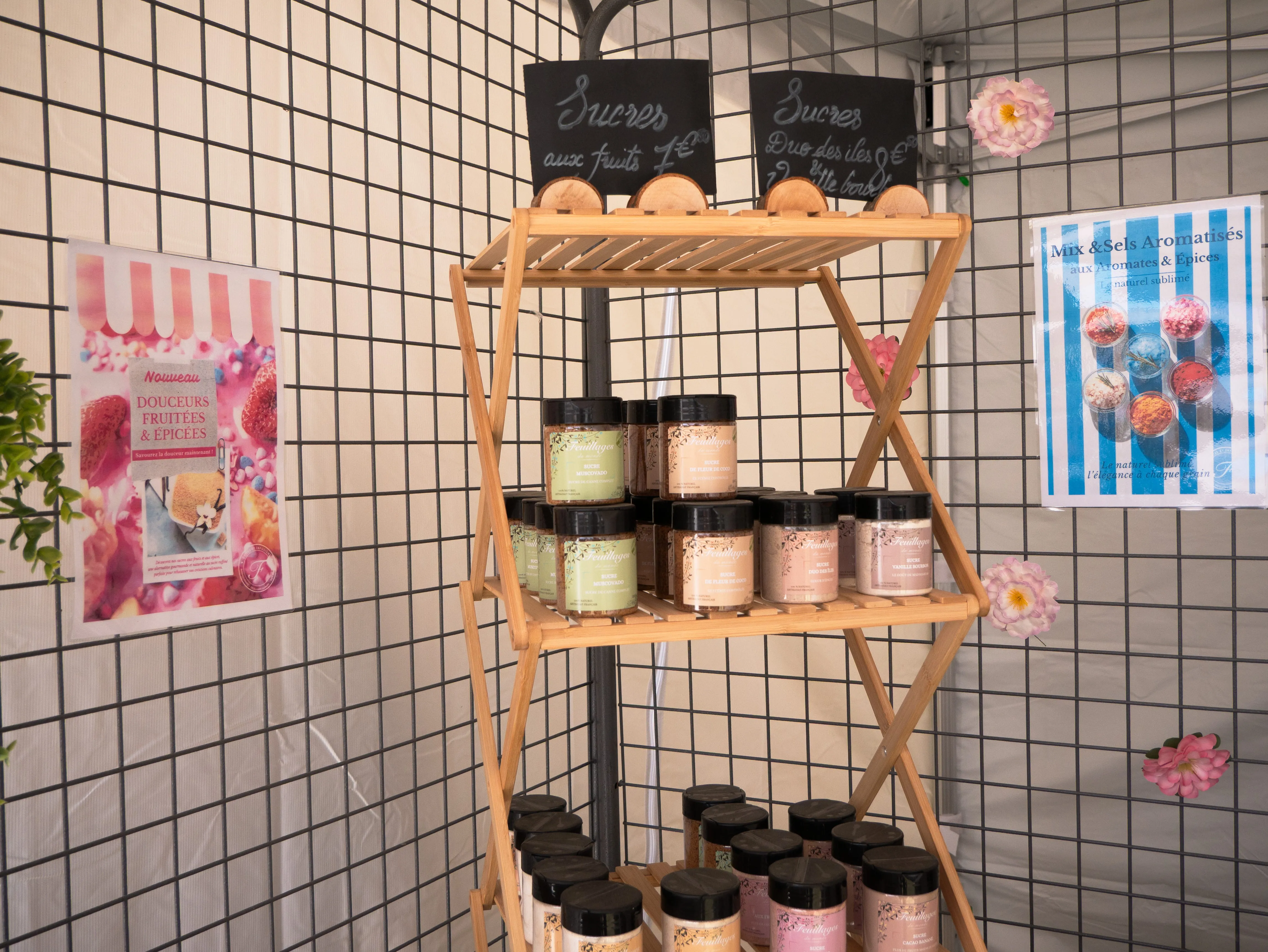 Étagère en bois avec pots de sucre aromatisé, affiches colorées et fleurs sur un grillage.