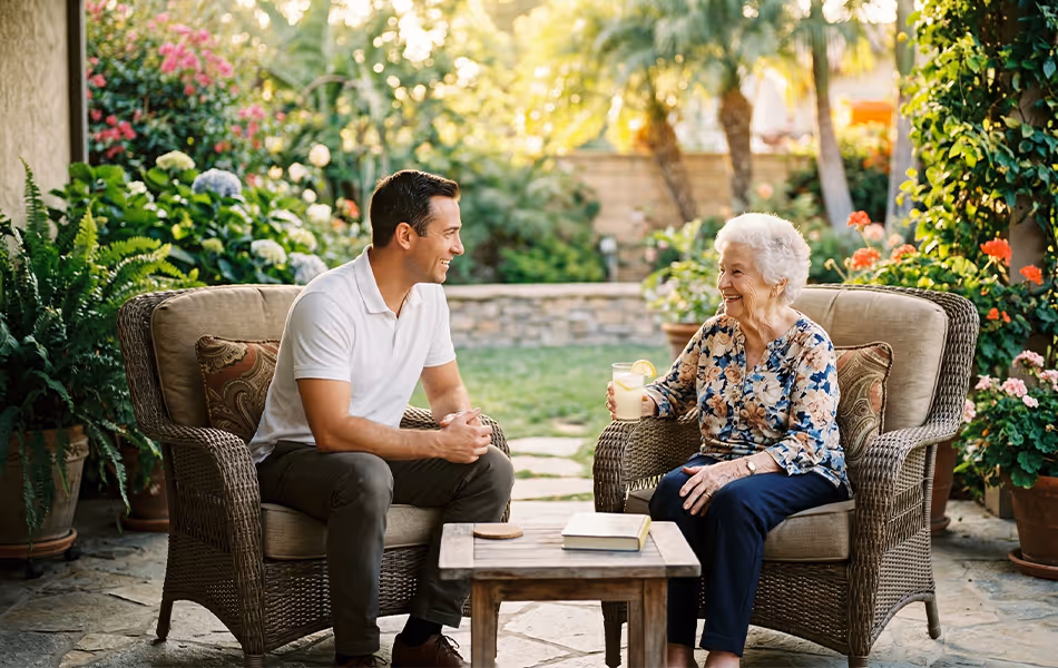 An elderly woman and a younger man smiling and talking while sitting on wicker chairs in a garden patio with a wooden table between them.