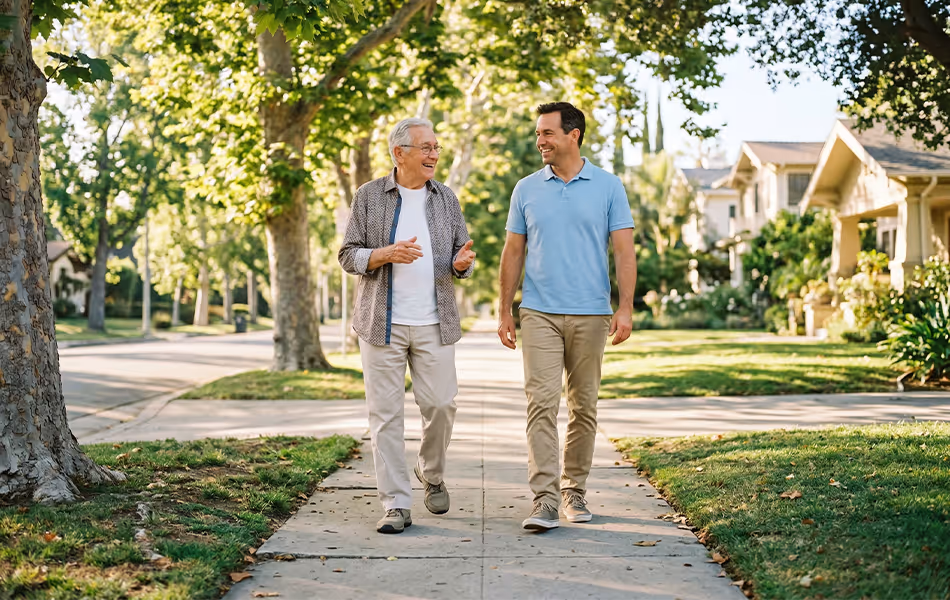 An elderly man and a younger man smiling and walking together down a tree-lined suburban sidewalk on a sunny day.