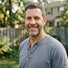 Smiling middle-aged man with short hair and beard wearing a gray shirt outdoors with greenery and fence in the background.