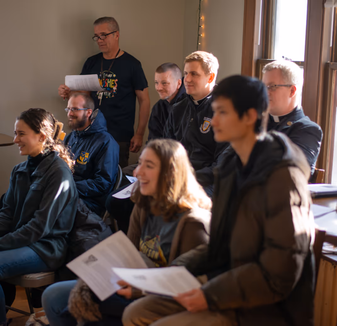 Group of people sitting indoors, some holding papers, smiling and engaging in a discussion near a window with natural light.