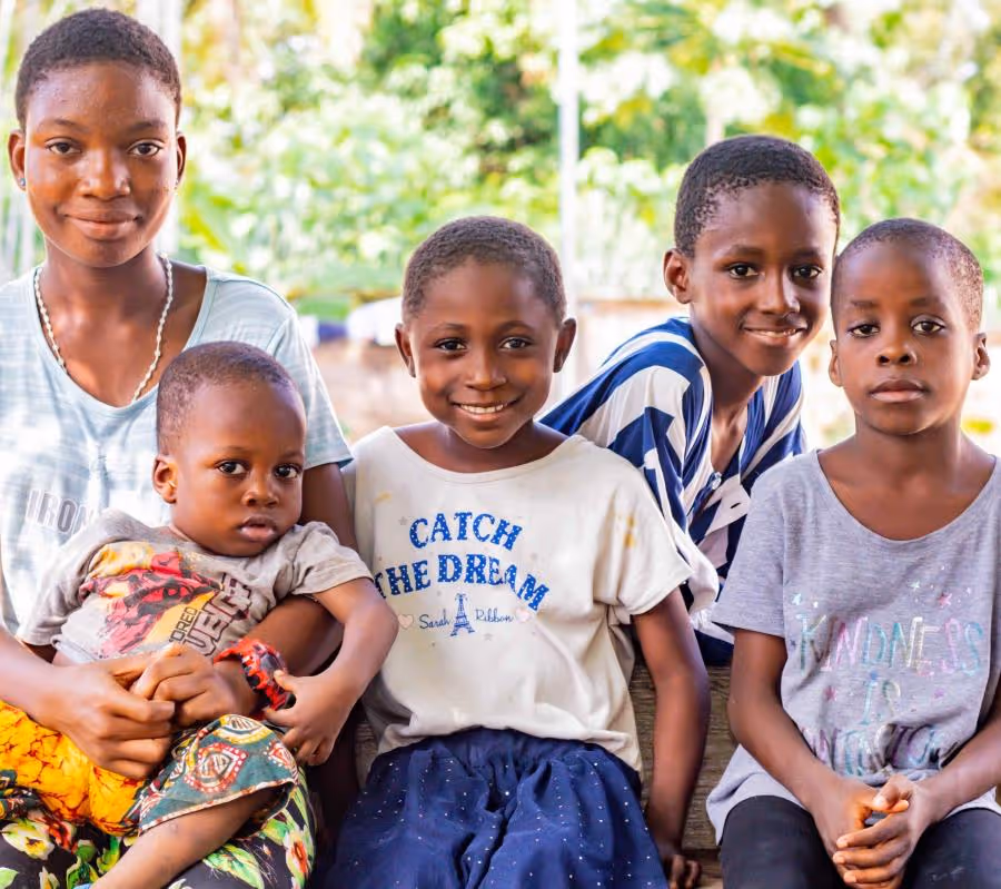 Five smiling Nigerian children sitting together outdoors with green foliage in the background.