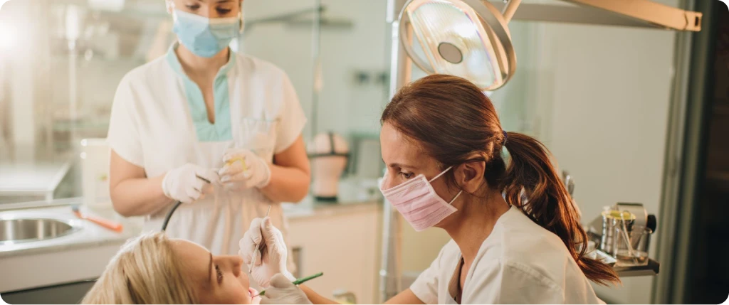 Dentist wearing a pink mask examining a female patient's teeth while an assistant in a blue mask prepares tools in the background.
