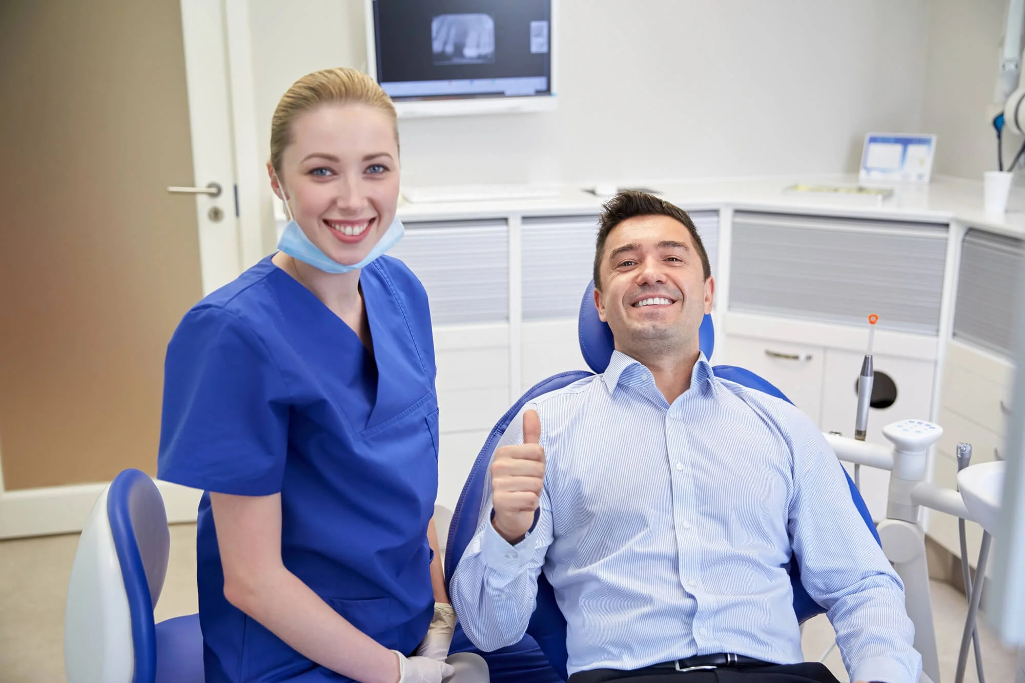 Smiling male dental patient in chair giving thumbs up next to female dentist in blue scrubs in a clinic.