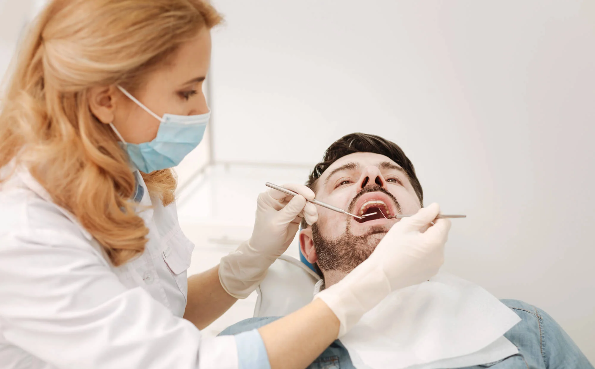 Dentist wearing a mask and gloves examining a male patient's open mouth using dental tools.