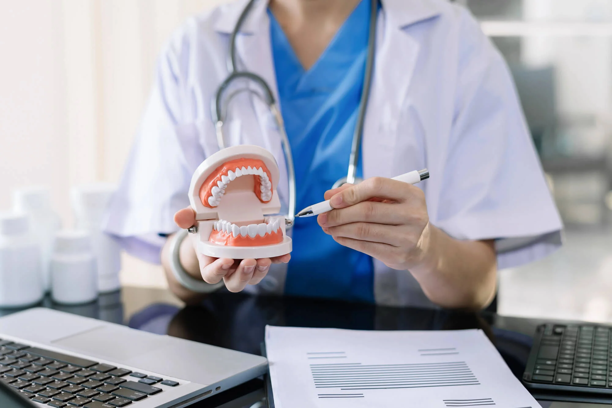 A healthcare professional wearing a lab coat and stethoscope holding a dental model and pointing at it with a pen.