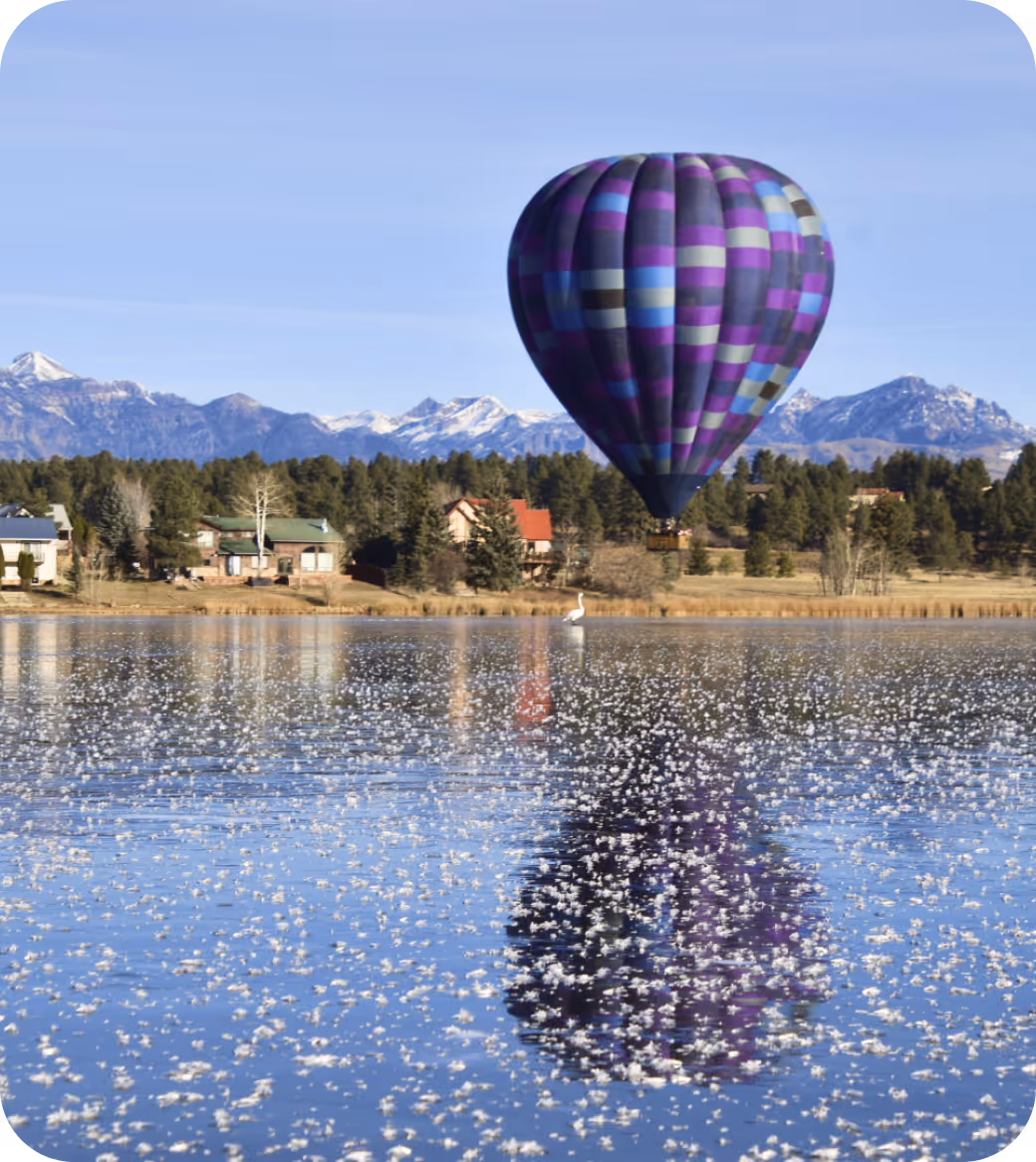 Purple and blue checkered hot air balloon reflected on a lake with snow-capped mountains and houses in the background.