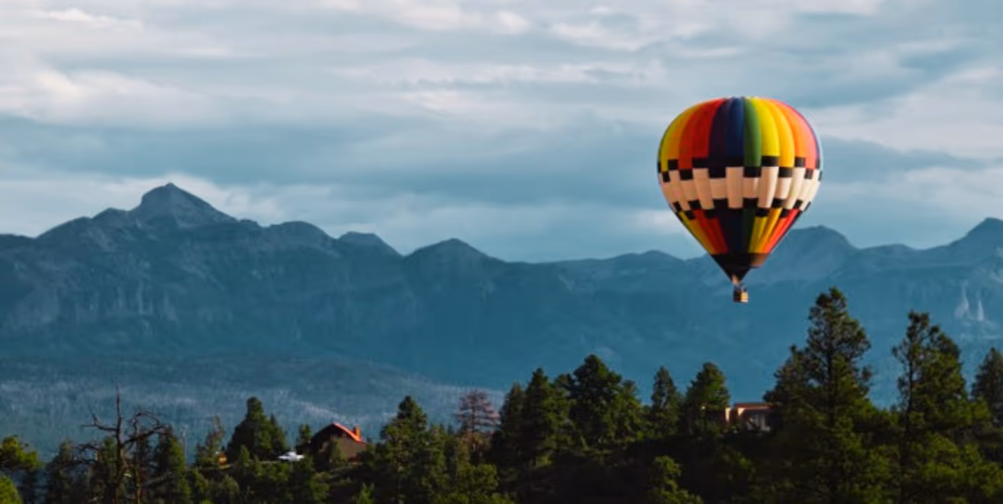 Colorful hot air balloon floating over forested hills with mountains in the background under a cloudy sky.
