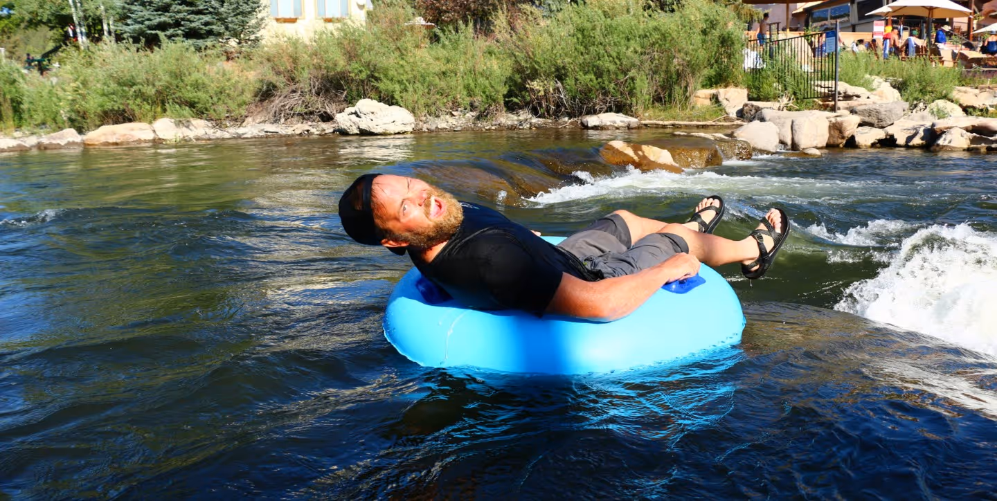Man wearing a black shirt and cap floating on a blue inner tube in a river with greenery and rocks in the background.