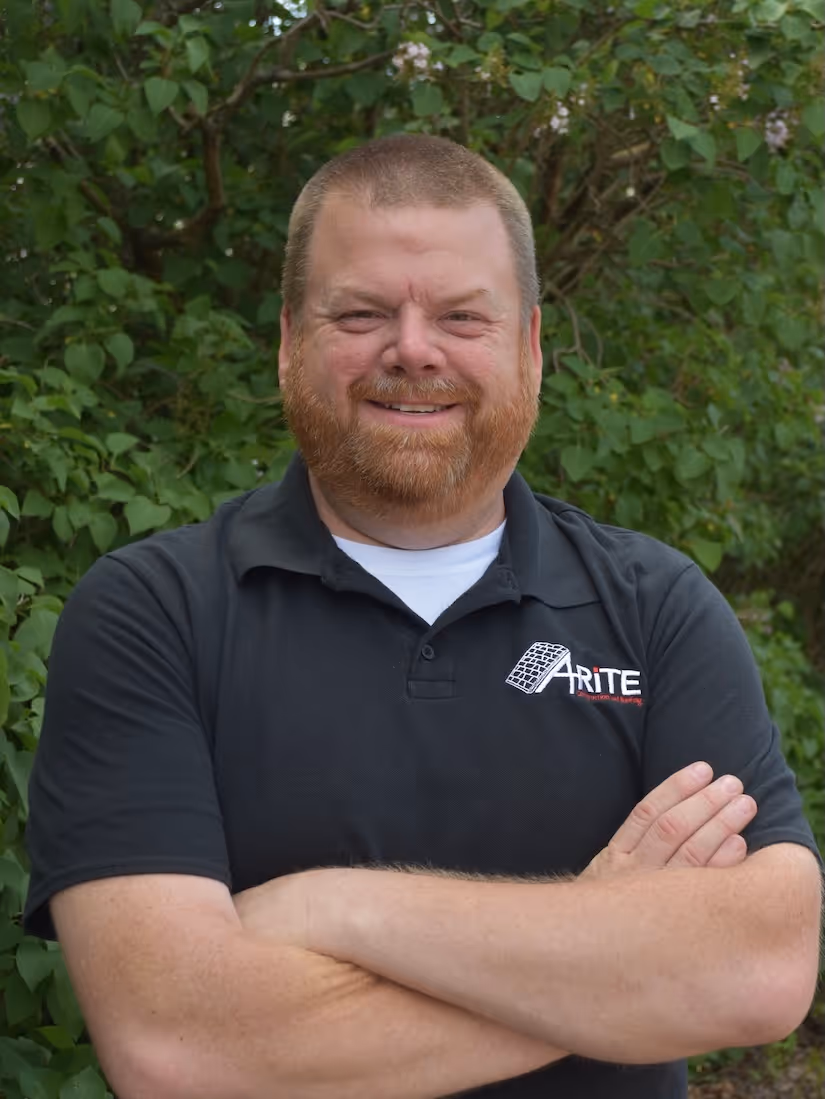 Smiling man with a beard wearing a black polo shirt with a white logo, standing with arms crossed in front of green foliage.