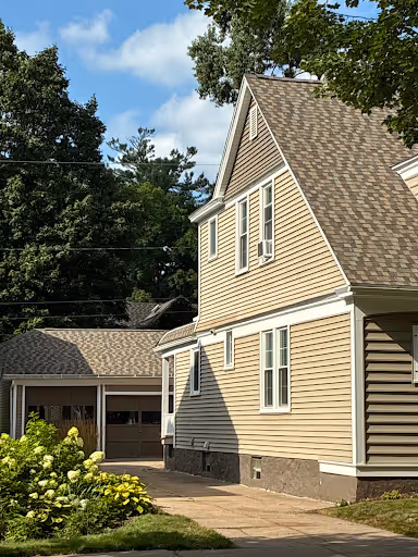 Side view of a beige two-story house with a steep roof, surrounded by green trees and a driveway leading to a garage.