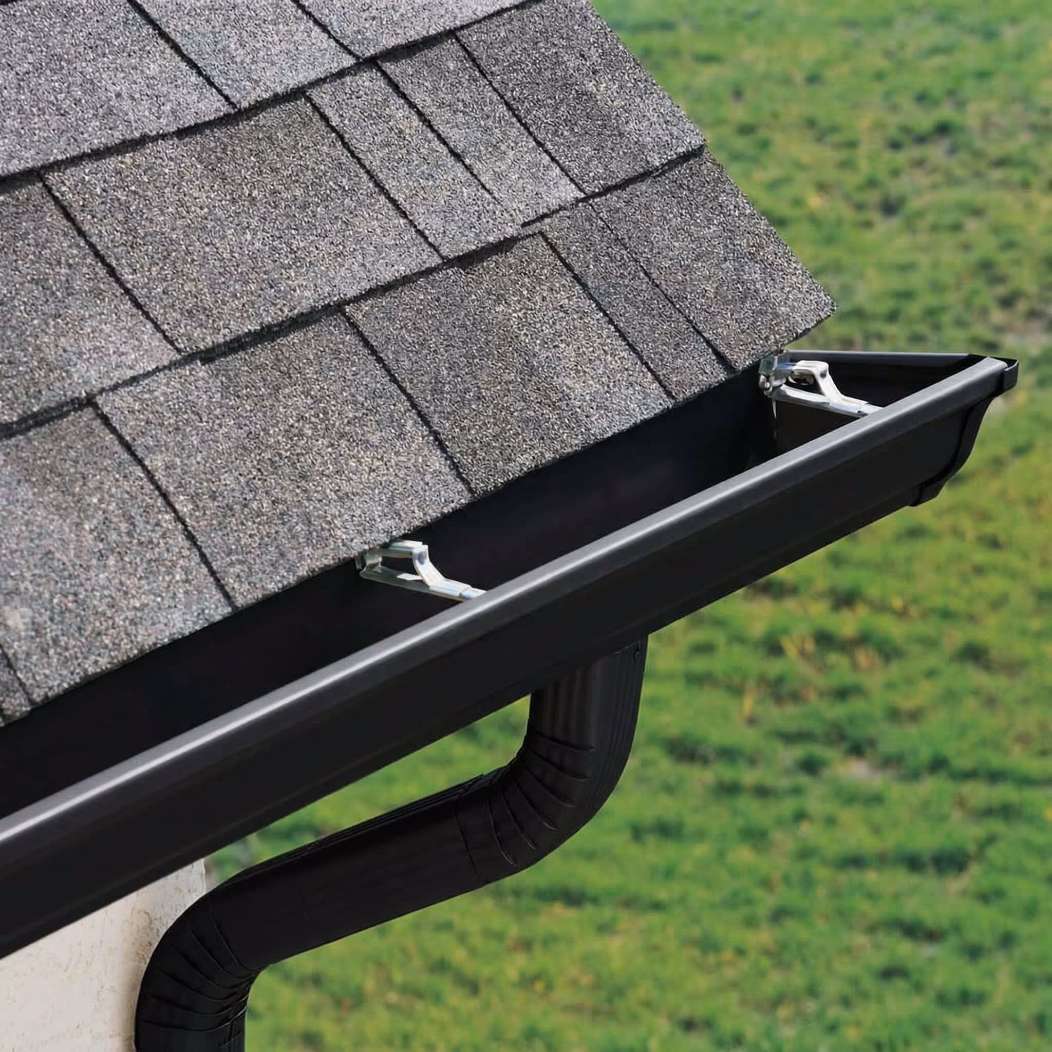Close-up of a black rain gutter and downspout attached to a grey shingled roof above a green grassy area.