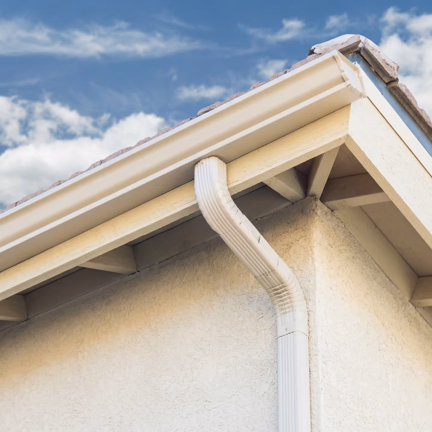 Corner of a house roof with white rain gutter and downspout against a partly cloudy blue sky.