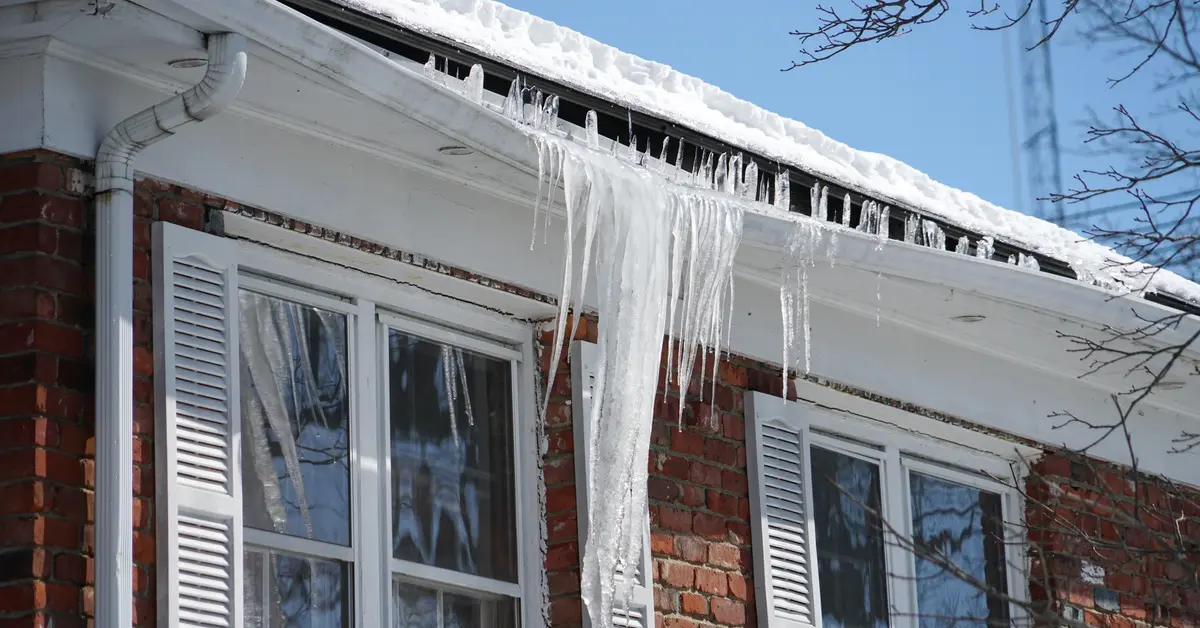 Large ice dam and icicles forming along roof edge and gutter on brick home in winter
