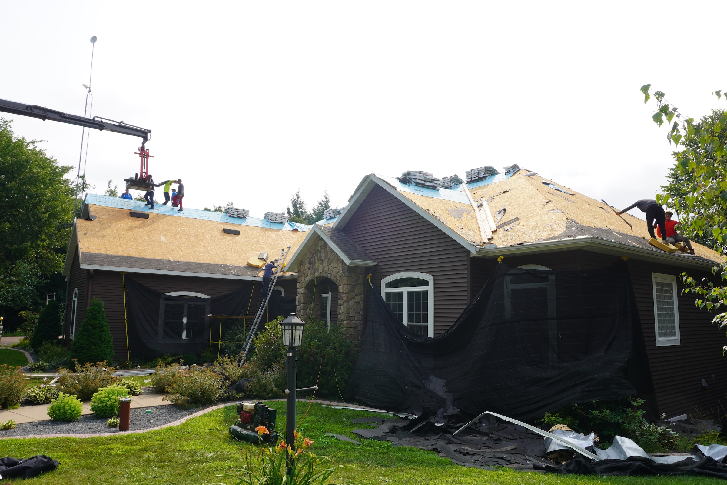 Front view of the same home during roof replacement, with old shingles removed, underlayment installed, workers on the roof, and protective coverings around the walls and landscaping.