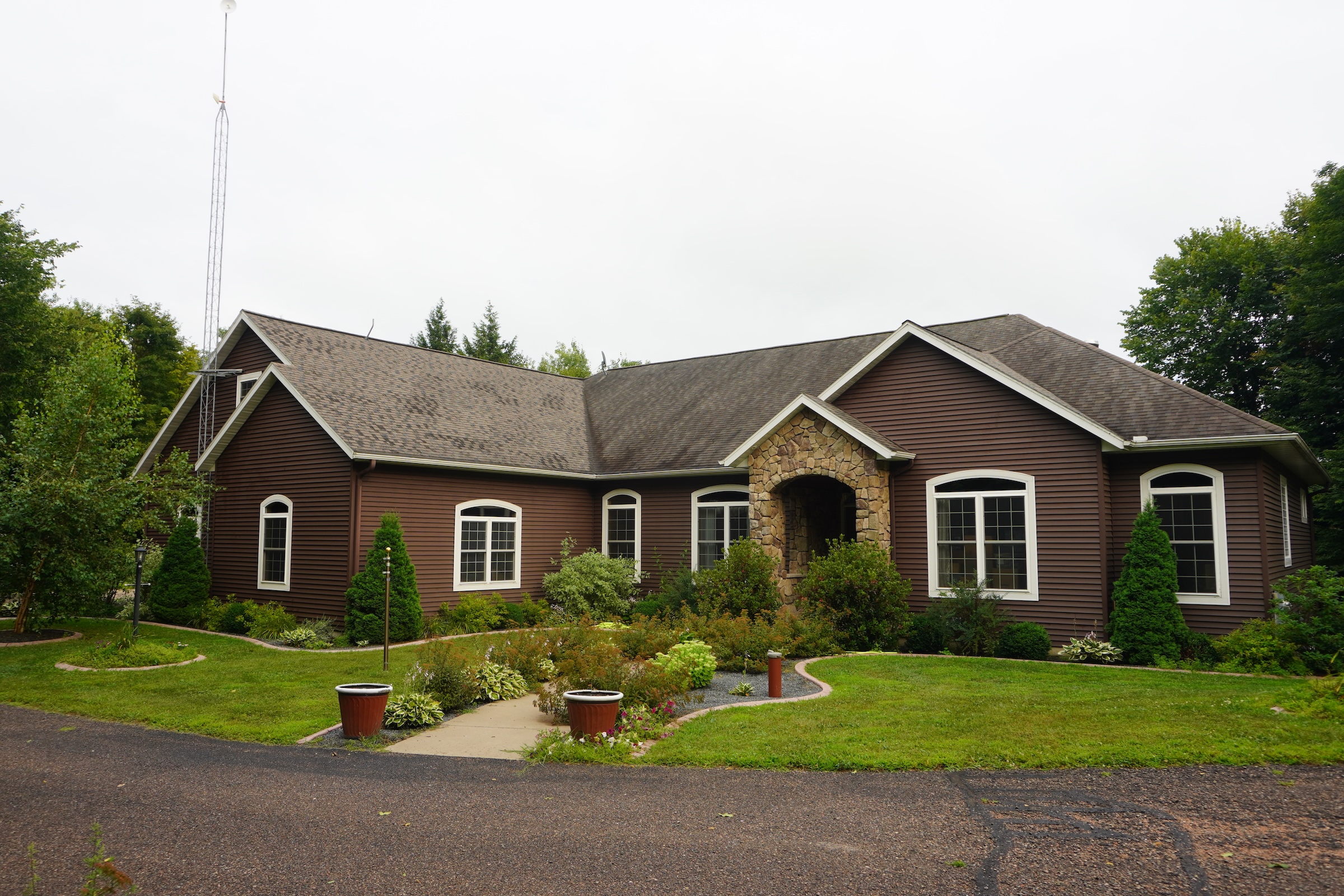 Front view of a large brown house with a stone entryway and an older shingle roof before replacement, surrounded by landscaped lawn and shrubs.