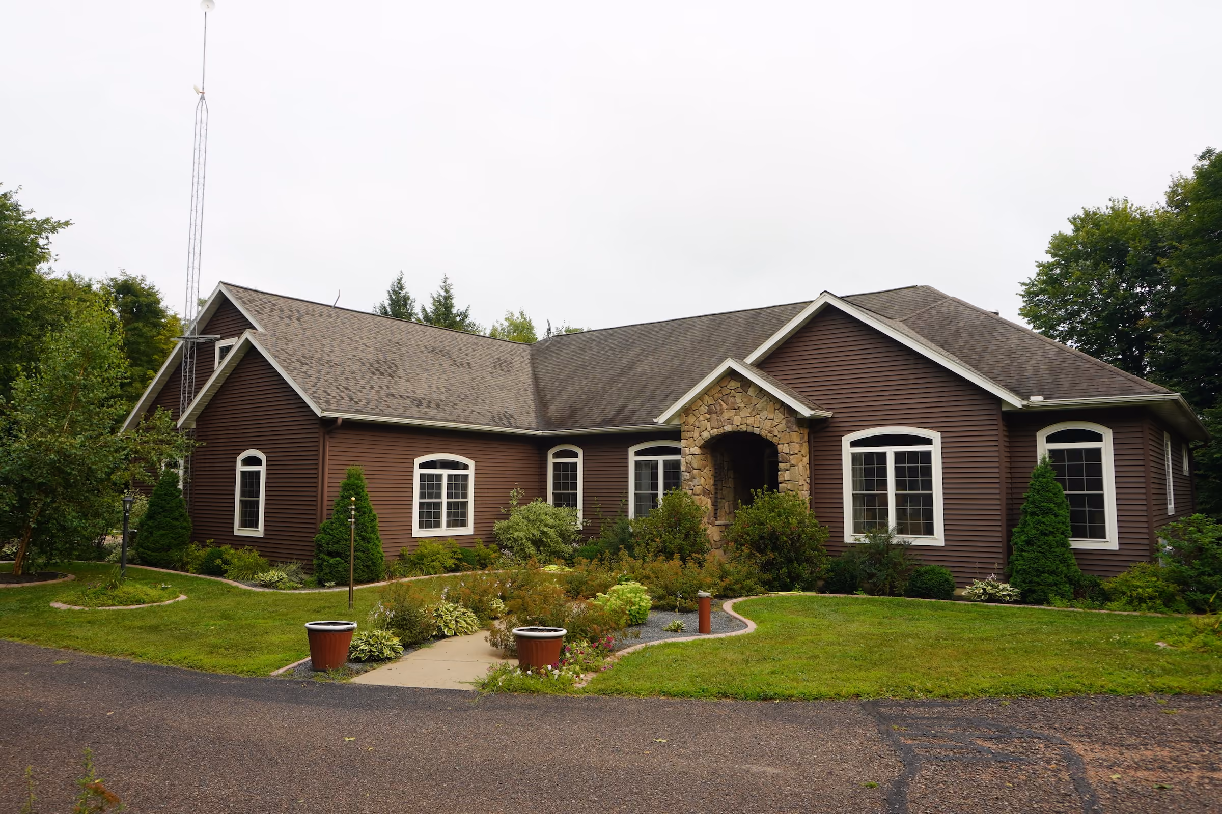 Front view of a large brown house with a stone entryway and an older shingle roof before replacement, surrounded by landscaped lawn and shrubs.