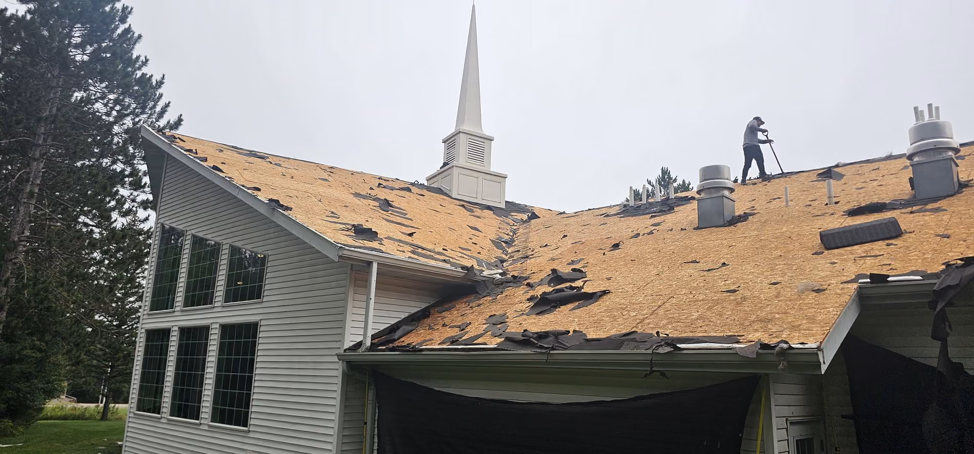 Large church roof in Merrill, WI during tear-off with wind-damaged shingles removed and roof decking exposed.